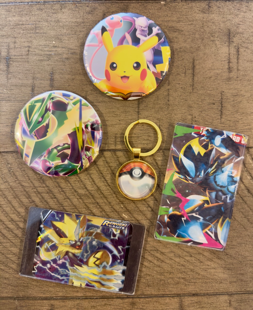 Katie’s handmade Pokémon pieces on a wooden table: two round designs, two rectangular card designs, and a gold-tone Poké Ball keychain.